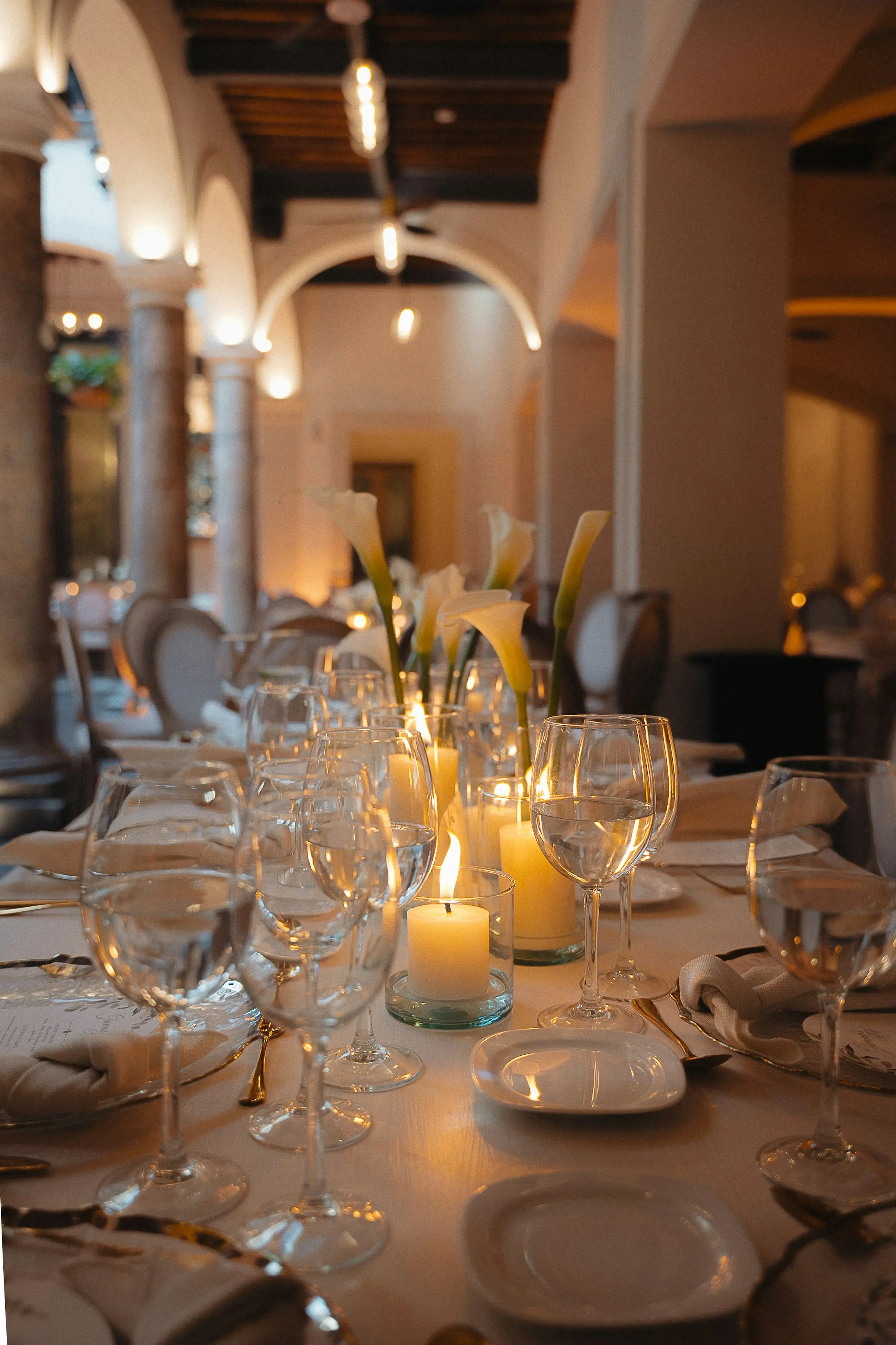 Table dressée à la lueur des bougies : verres à pied, assiettes blanches et arums blancs, sous des arcades éclairées