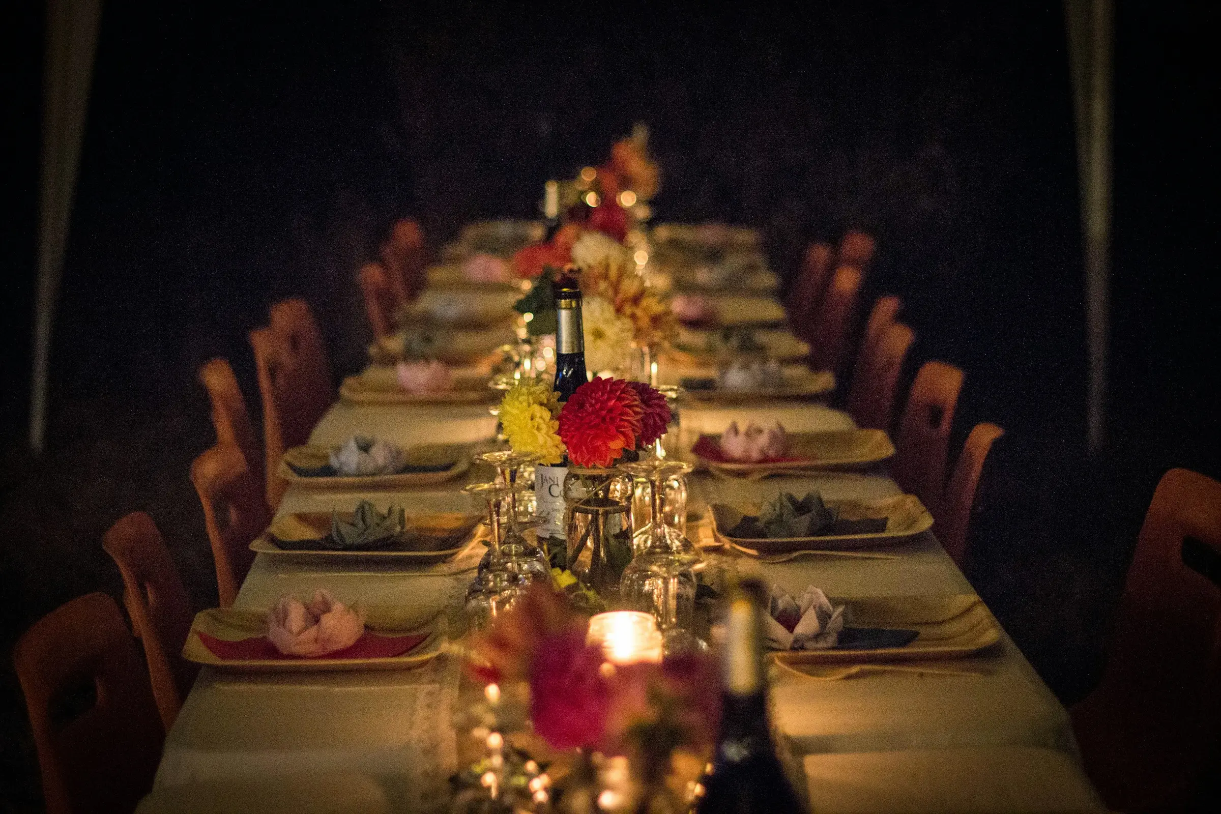 Longue table dressée dans la pénombre, éclairée à la bougie, bouquets de dahlias et serviettes pliées en fleur