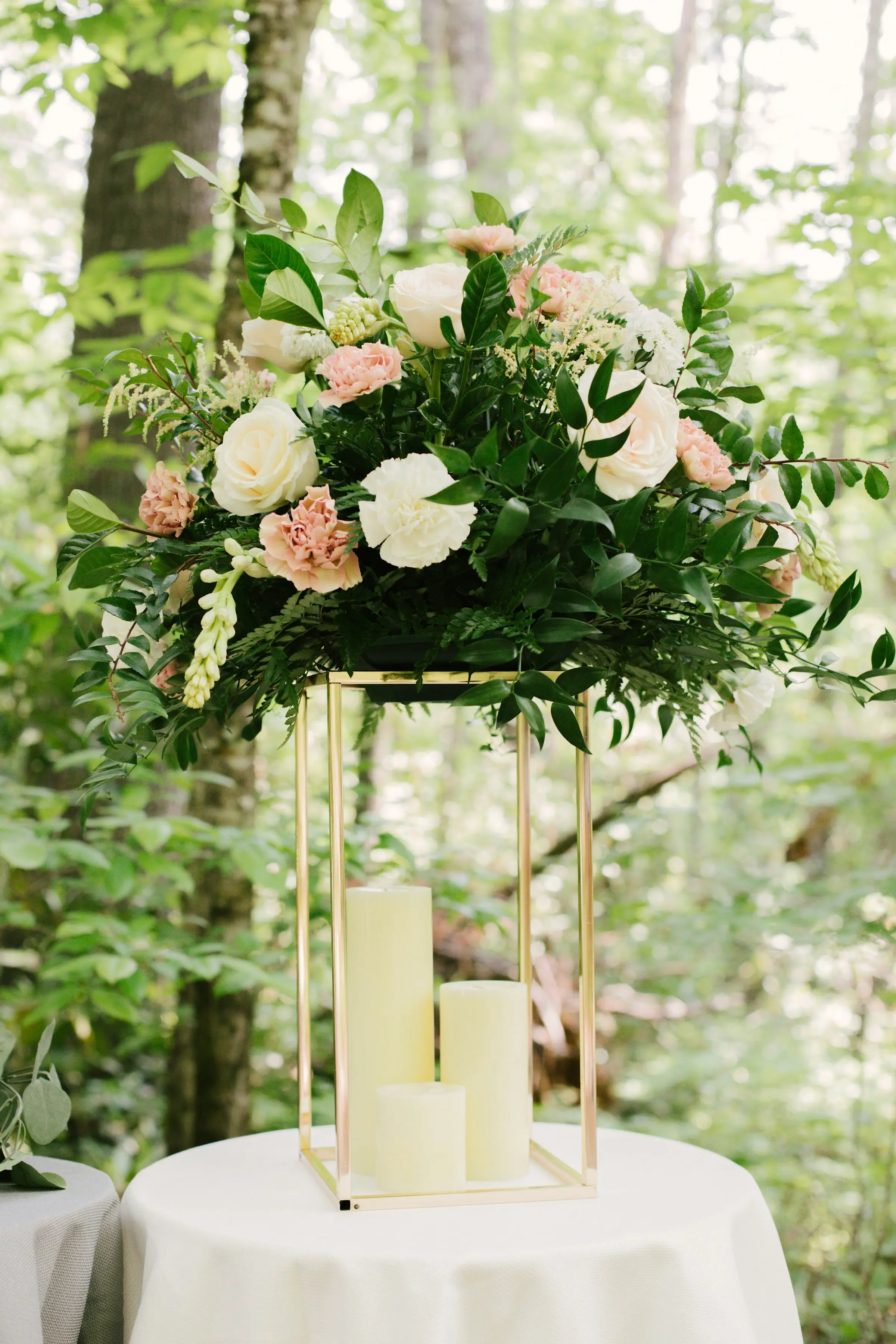Composition de roses blanches et poudrées sur une colonne dorée et bougies, dans un sous-bois