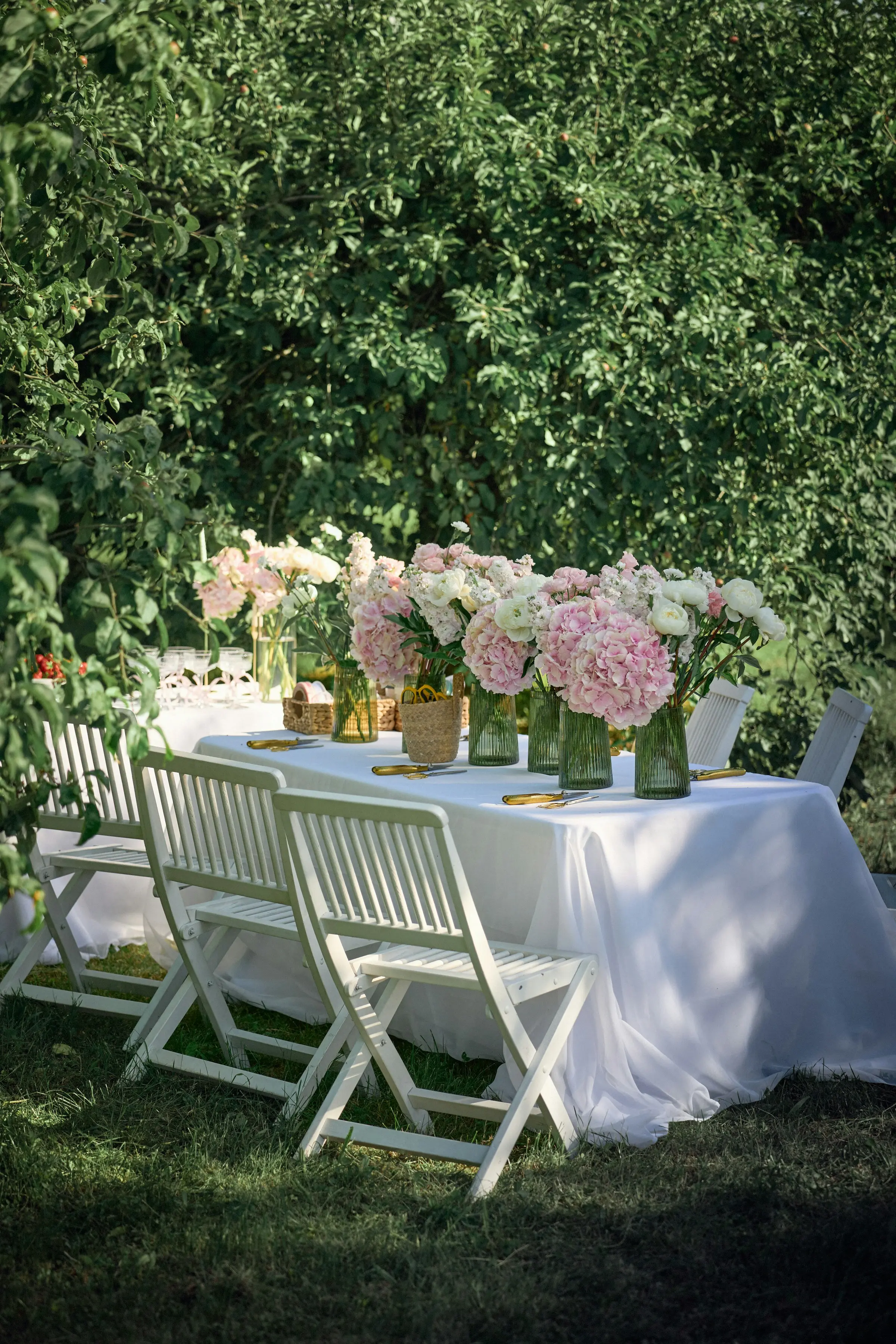 Table nappée de blanc décorée d’hortensias roses, chaises pliantes blanches dans un jardin