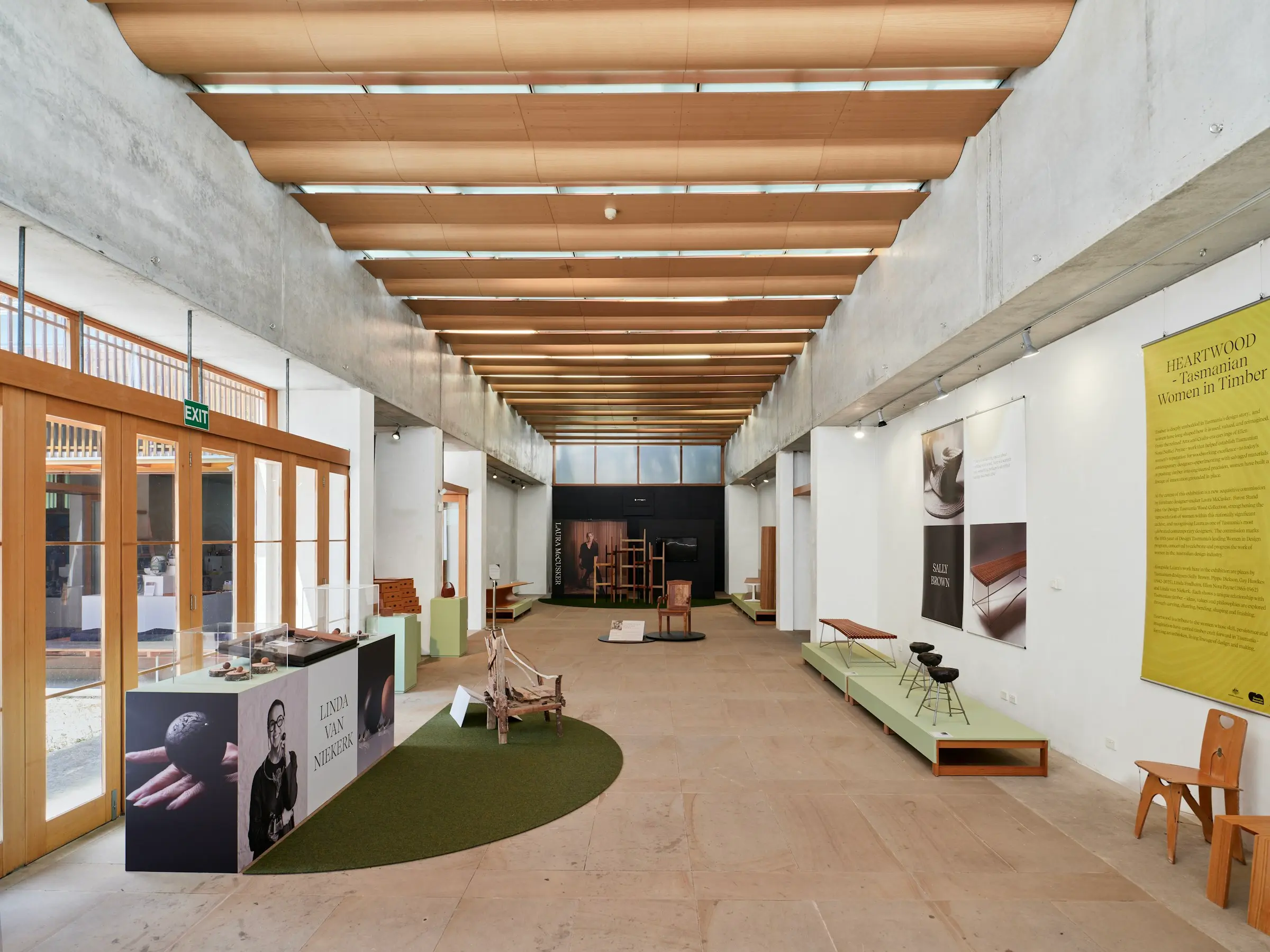 Galerie d’exposition au plafond de lattes de bois, vitrines, mobilier exposé et panneaux le long d’une allée