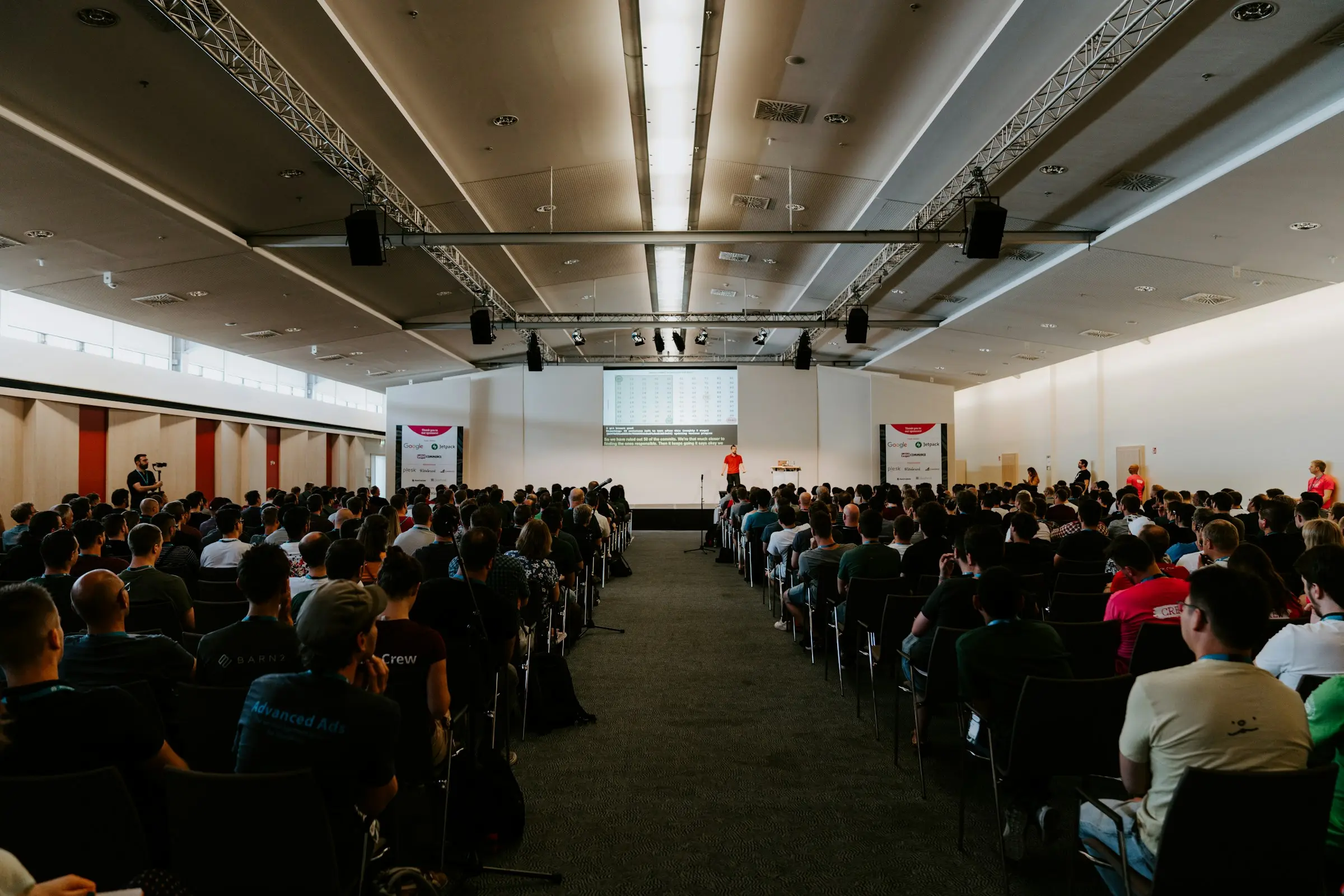Vaste salle de conférence comble, public de dos face à une scène lointaine sous des structures d’éclairage