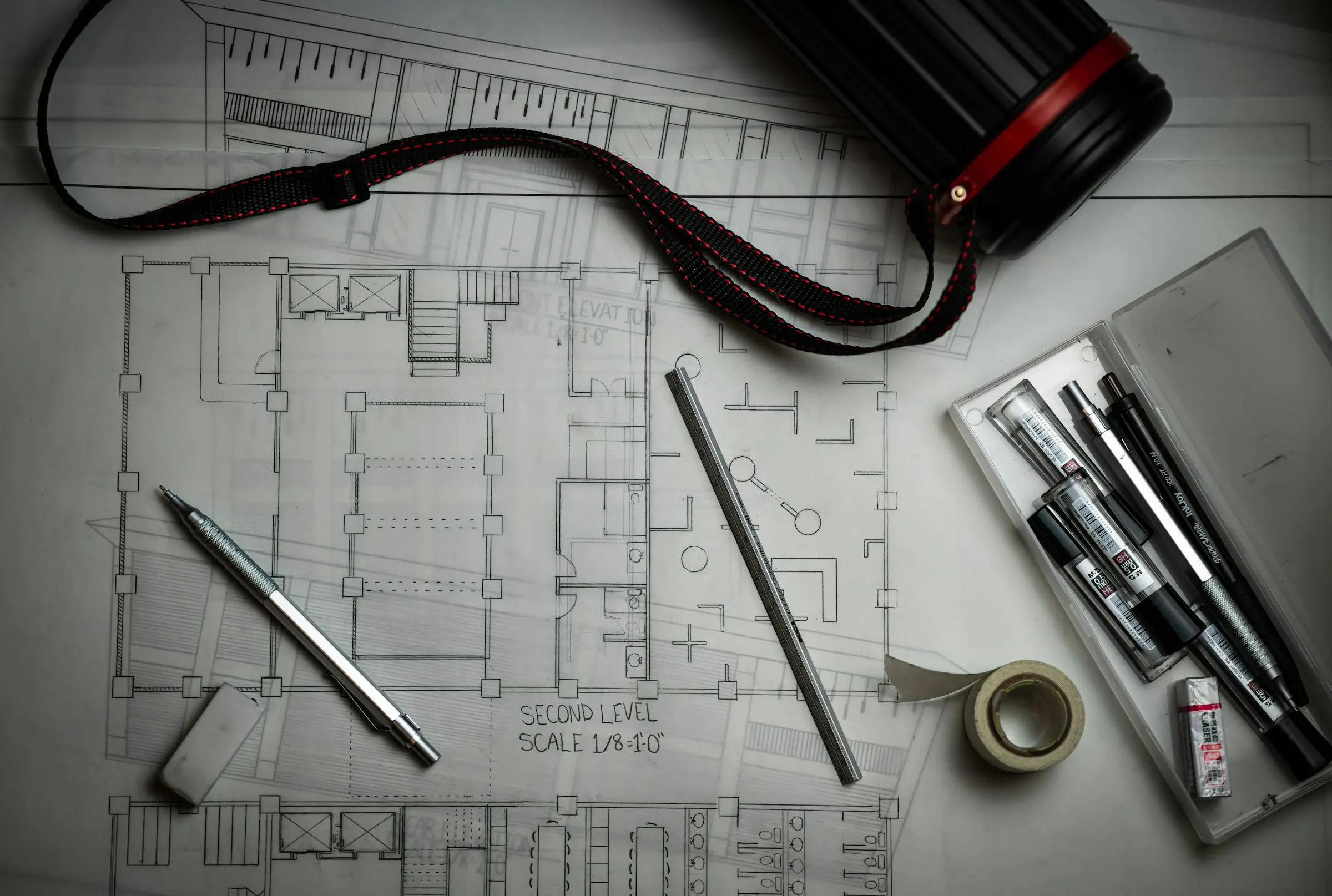 Plan d’architecte annoté, crayons, réglet, ruban adhésif et tube à plans sur une table de travail