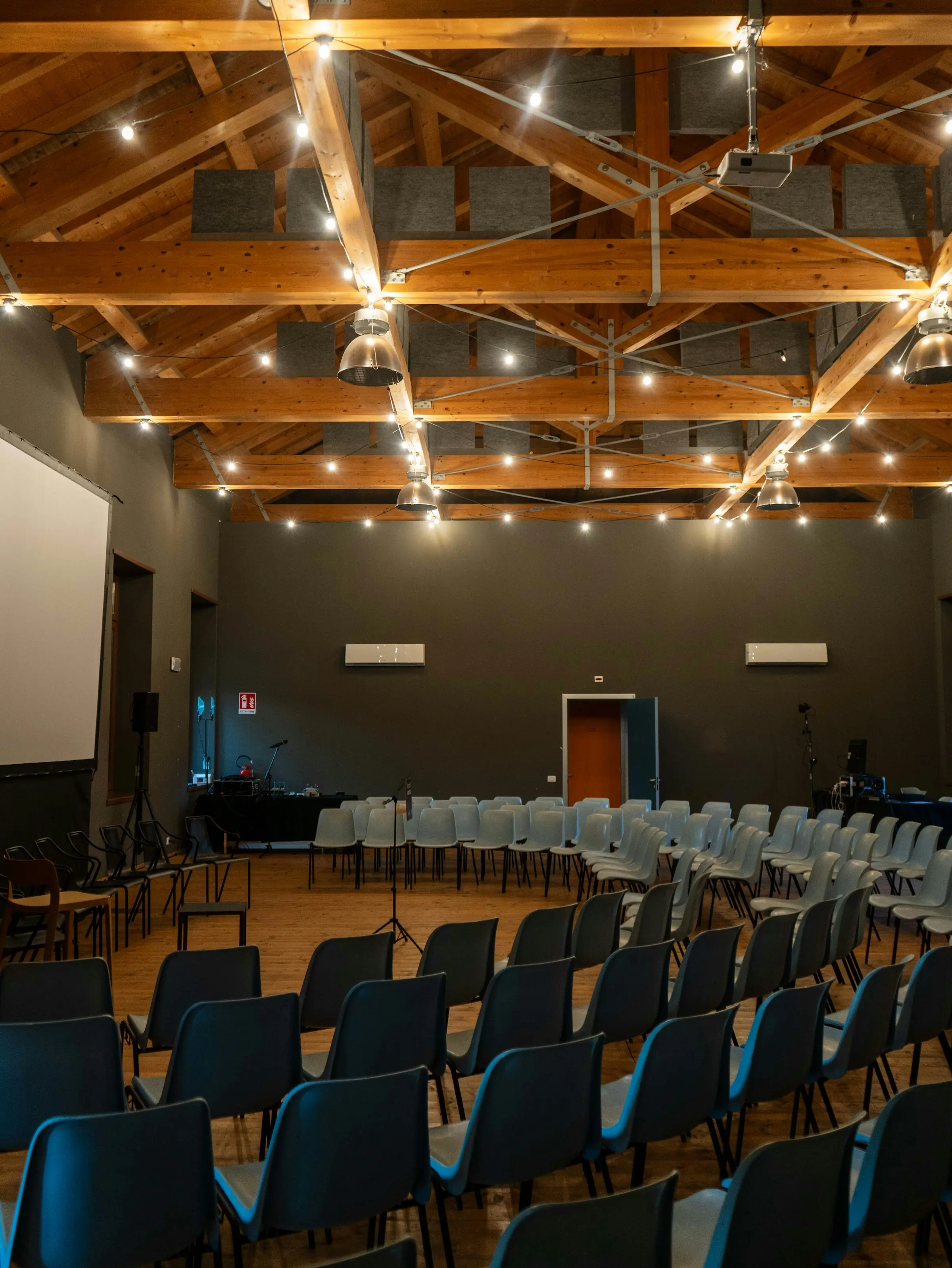 Salle polyvalente sous charpente en bois et guirlandes lumineuses, chaises disposées en arc de cercle avant l’arrivée du public